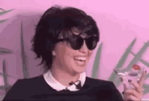 a woman wearing sunglasses is sitting in front of a pink wall holding a martini .