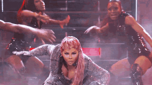 a woman with pink hair is kneeling down on a stage surrounded by dancers .