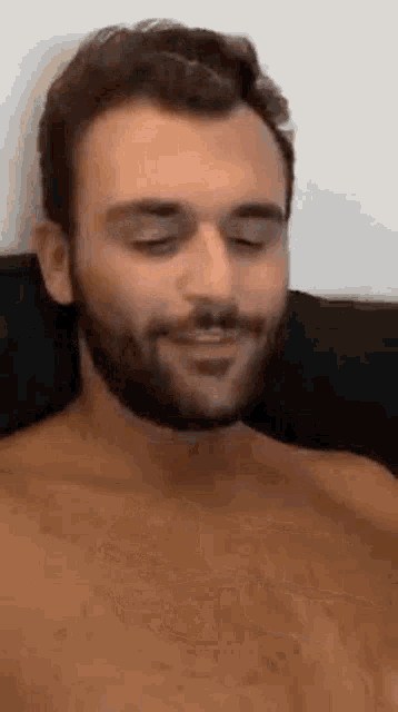 a shirtless man with a beard is smiling while sitting on a couch .