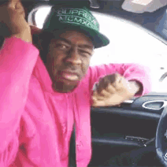a man wearing a pink hoodie and a green hat with the word supra on it is sitting in a car