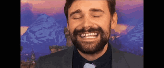 a man with a beard is smiling with his eyes closed in front of a mountain