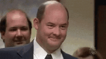 a bald man in a suit and tie is smiling in front of another bald man .