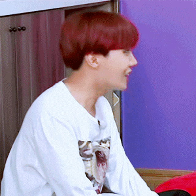 a young man with red hair is wearing a white shirt and sitting in front of a purple wall .