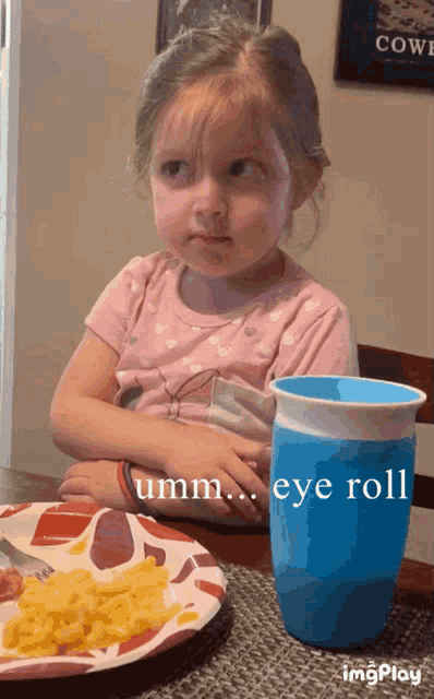 a little girl is sitting at a table with a plate of food and a cup that says eye roll