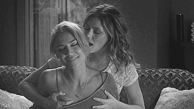 two women are sitting on a couch and one is kissing the other on the chest .