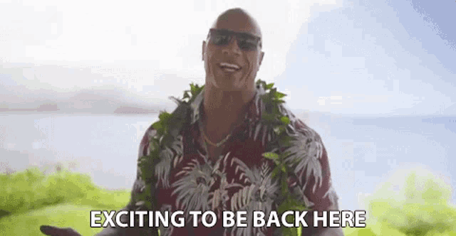 a man wearing a lei and sunglasses is standing in front of the ocean and saying `` exciting to be back here '' .