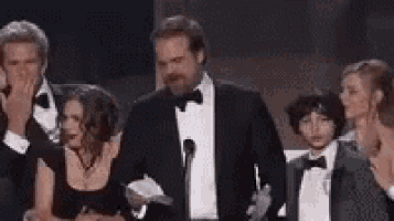 a man in a tuxedo is standing in front of a microphone with a group of people behind him .