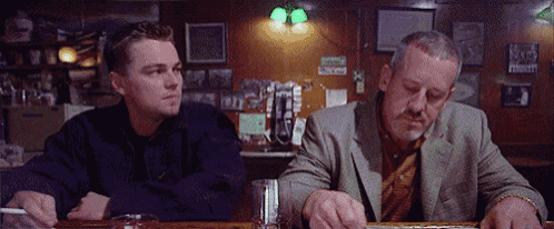 two men are sitting at a bar one is smoking a cigarette and the other is drinking a beer