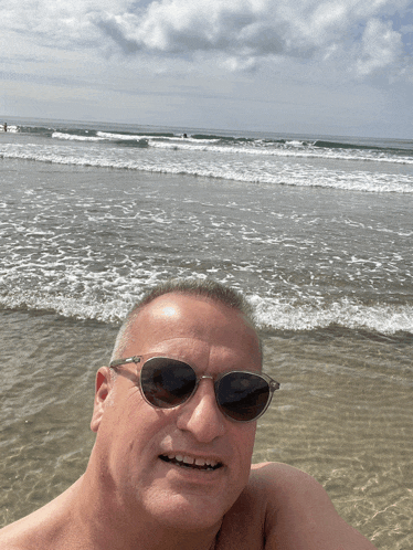 a shirtless man wearing sunglasses takes a picture of himself on the beach