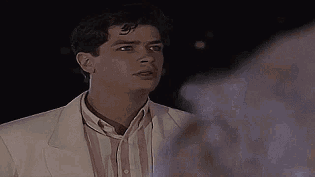 a man in a white jacket and pink and white striped shirt stands in front of a fountain