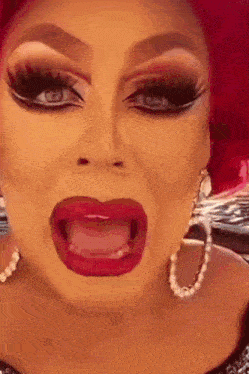 a drag queen with red hair and pearl hoop earrings is making a funny face with her mouth open .