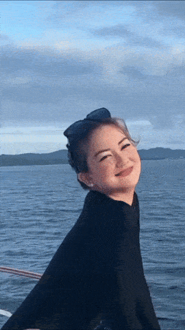 a woman wearing sunglasses and a black sweater smiles in front of a body of water