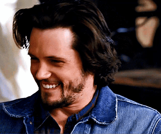 a man with long hair and a beard wearing a denim jacket smiles