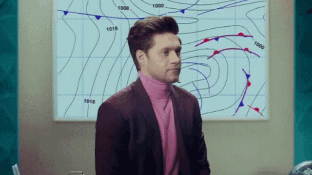 a man wearing a pink turtleneck and a suit is standing in front of a weather map .