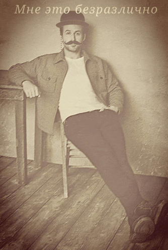 a man with a hat and mustache is sitting on a chair with his legs crossed in front of a sign that says mne