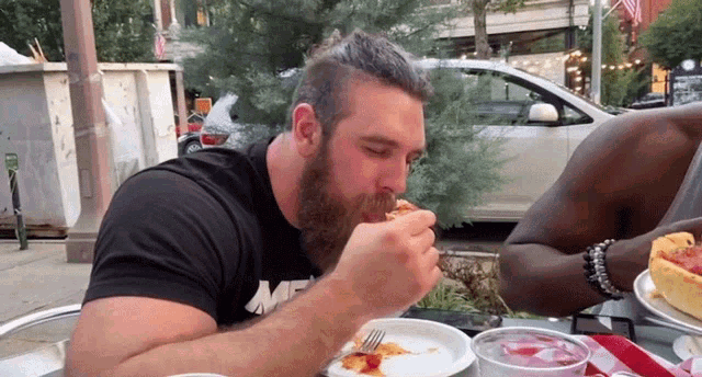a man with a beard is eating a slice of pizza while wearing a black shirt with the letter m on it