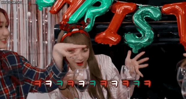 two girls are sitting at a table with balloons in the background and one girl is wearing a christmas headband .