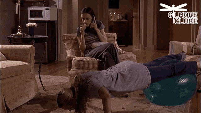 a woman is doing push ups while another woman sits in a chair and talks on a cell phone