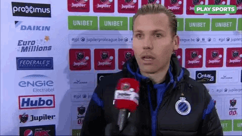 a man is talking into a microphone in front of a wall with advertisements for jupiter
