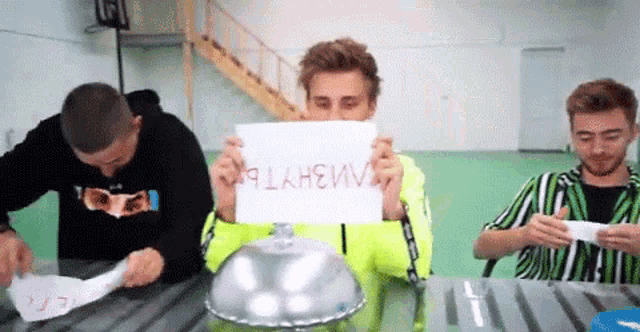 three men are sitting at a table holding a sign in front of their face .