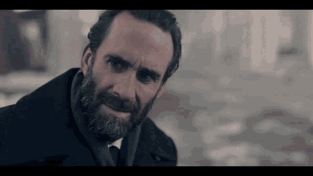 a man with a beard wearing a black coat and tie looks at the camera
