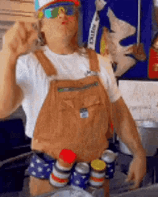 a man wearing overalls , a hat , sunglasses and a belt of cans is standing in a kitchen .