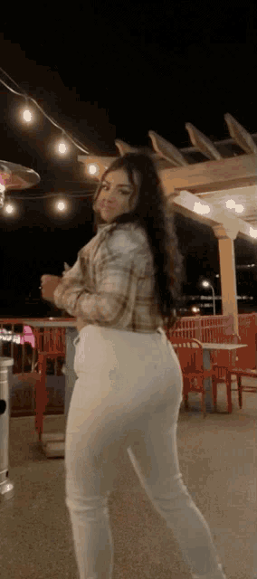 a woman wearing white pants and a plaid shirt is standing in front of a restaurant at night .