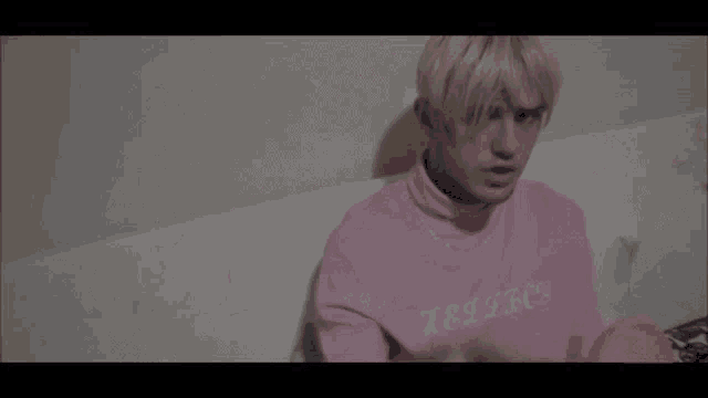 a man with pink hair is sitting on a bed wearing a pink sweatshirt and smoking a cigarette .