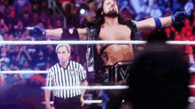 a wrestler is standing in a wrestling ring with his arms outstretched while a referee watches .