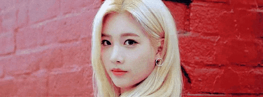 a woman with blonde hair is standing in front of a red brick wall and looking at the camera .