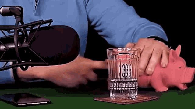 a man is sitting at a table with a microphone and a glass of water next to a pink piggy bank .