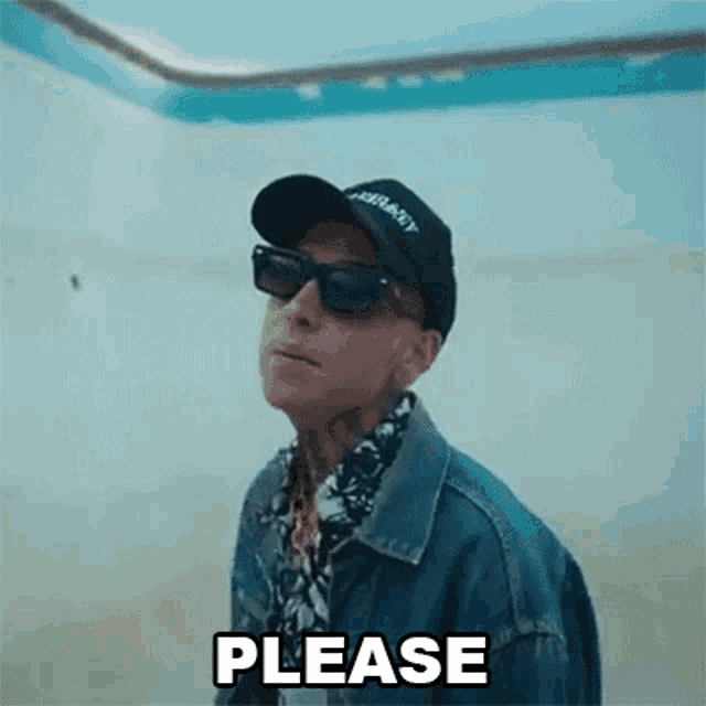 a man wearing sunglasses , a hat and a denim jacket is standing in a room and saying please .