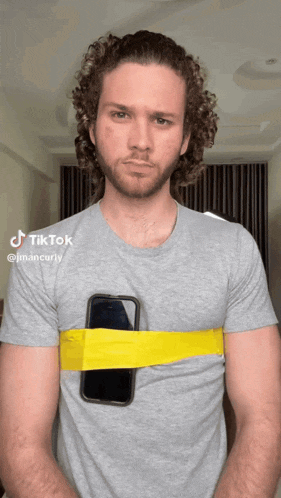 a man with curly hair is wearing a grey shirt with a yellow rubber band around his chest holding a cell phone
