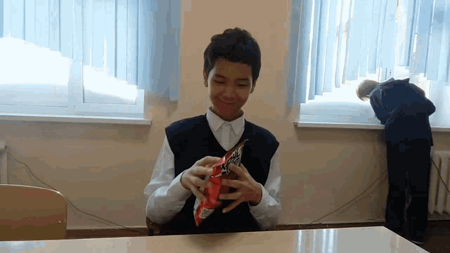 a boy is sitting at a desk holding a bag of fritos