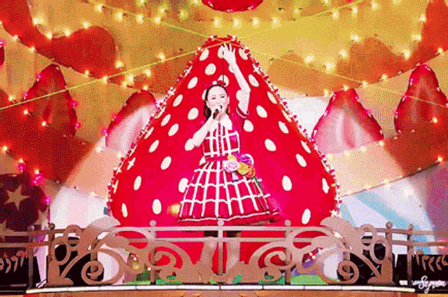 a girl in a red dress is singing into a microphone in front of a giant strawberry shaped decoration .