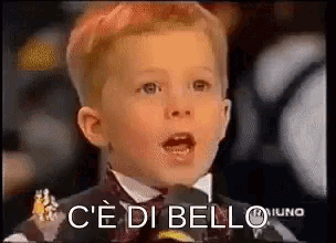a young boy in a suit and bow tie is speaking into a microphone with the words c'e di bello behind him