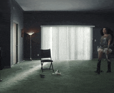 a woman stands in a dark room with a chair and a lamp