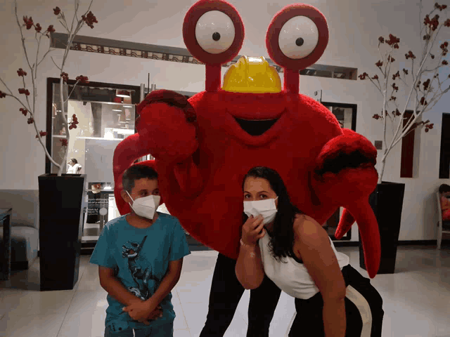 a woman and a boy wearing face masks pose with a red crab mascot