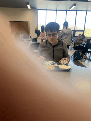 a boy wearing glasses sits at a table with a plate of food in front of him