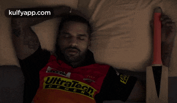 a man laying on a bed with a cricket bat and a ultratech shirt on