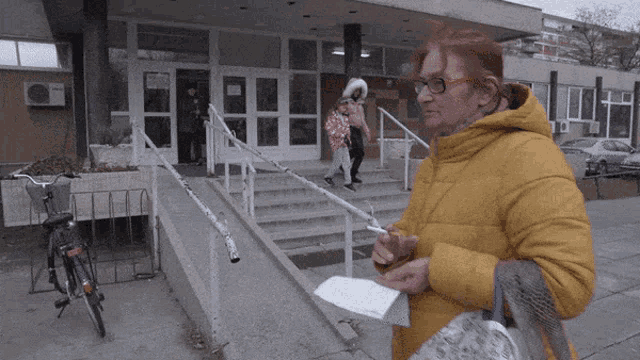 a woman in a yellow jacket is smoking a cigarette in front of a building
