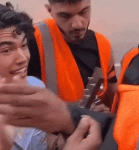 a man in an orange vest is holding a guitar while another man looks on .