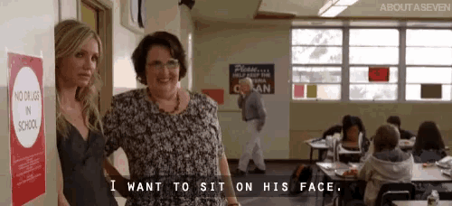 two women are standing next to each other in a school hallway and one of them is saying i want to sit on his face .