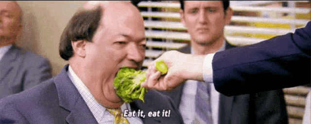 a man in a suit and tie is eating broccoli while another man says " eat it eat it "