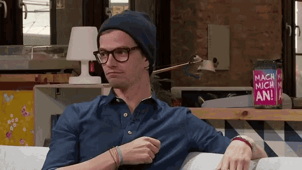 a man wearing glasses and a beanie sits on a couch next to a bottle of mach mich an