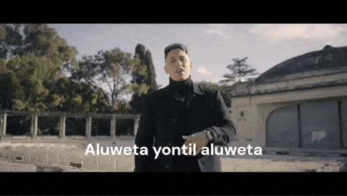 a man in a black coat stands in front of a building with the words aluweta yontil aluweta written below him