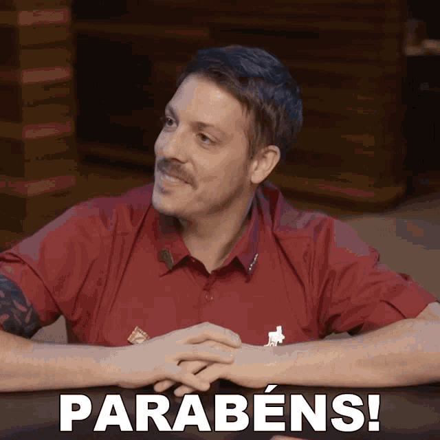 a man in a red shirt sits at a table with the words parabéns on the bottom