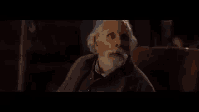 an elderly man with a beard is sitting in a chair with his mouth open in a dark room .