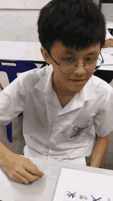 a young boy wearing glasses sits at a desk with a piece of paper that says ' x ' on it