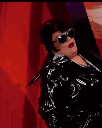 a drag queen is standing on a stage wearing a black jacket and a black dress .